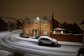 Suburban snow night This urban landscape photograph shows a suburban scene in Chesterfield, Derbyshire, United Kingdom during a snowy night in winter. The main subject is a red brick house typical of suburban architecture, illuminated by a streetlight that highlights the fresh layer of snow blanketing the ground, rooftops and parked car. The snow-covered street curves in front of the house, with tracks revealing recent travel through the suburb. Rows of houses in similar architectural style are visible in the background, contributing to the suburban setting commonly found in this part of the United Kingdom. The winter conditions are evident from the snow accumulation, and the nighttime setting is characterized by the warm glow from windows and streetlights, emphasizing the quiet atmosphere of a Derbyshire suburb.